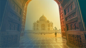 Blick durch einen aufwendig mit Schnitzereien verzierten Torbogen auf das Taj Mahal, Agar, mit seinem weiten Vorplatz bei Sonnenlicht.
