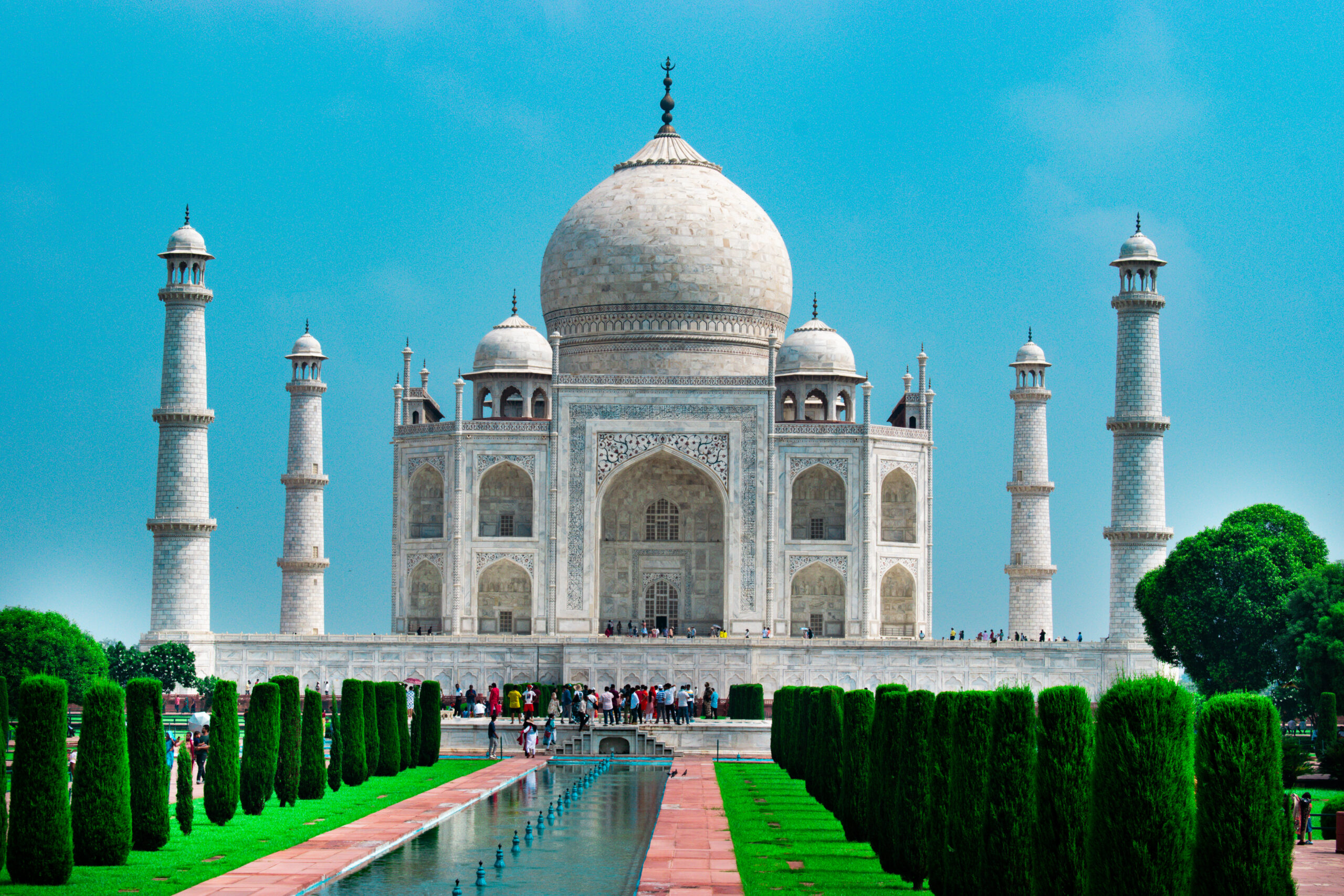 Vorderansicht des Taj Mahal in Agra vor hellblauem Himmel, im Vordergrund Menschen und ein blauer Wasserlauf, eingesäumt von grünen Büschen.