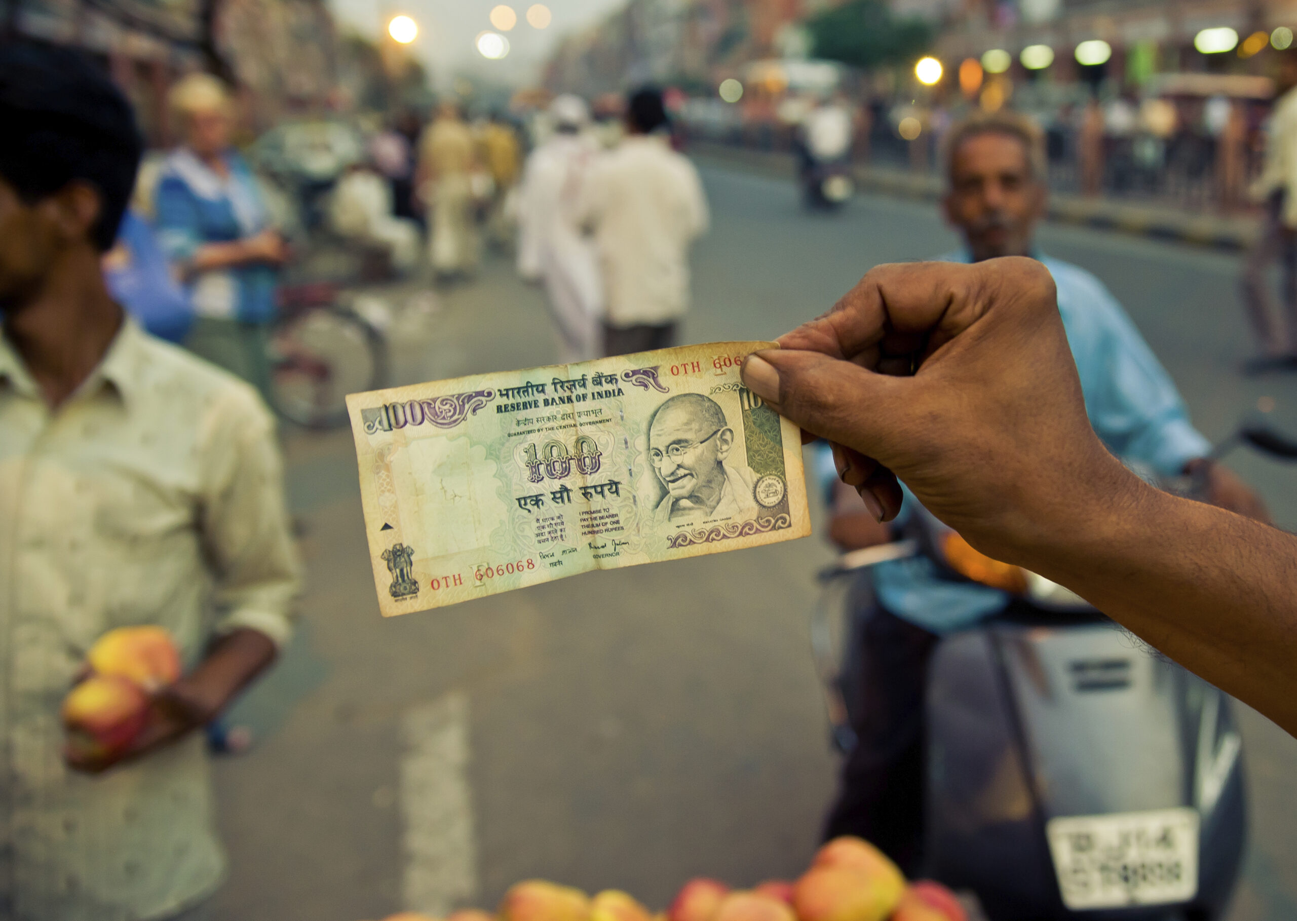 Eine Hand hält die Vorderseite einer 100 INR Banknote in die Kamera. Im Hintergrund verschwommen eine indische Straßenszene.