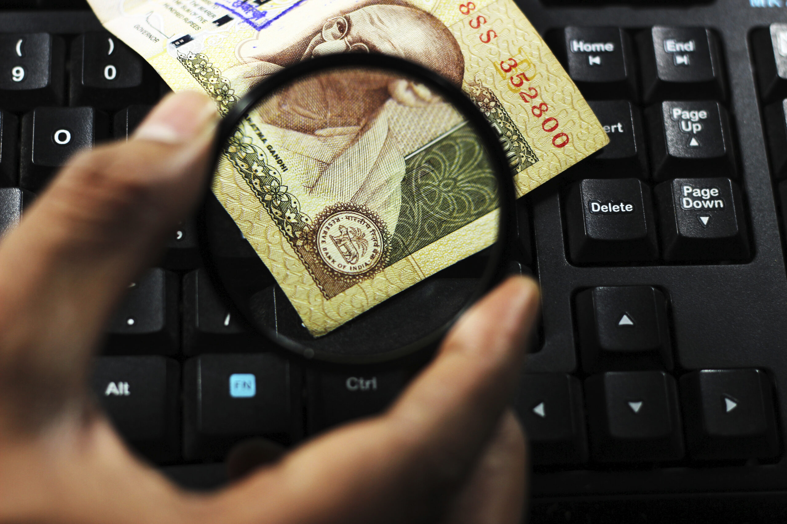 Der Bildausschnitt einer indische Banknote, die schräg auf dem Ausschnitt einer schwarzen Computertastatur liegt. Eine Hand hält eine Lupe über die rechte untere Ecke des Geldscheins und vergrößert so die Ansicht des RBI-Siegels mit Motiv des Ashoka-Löwenemblems.