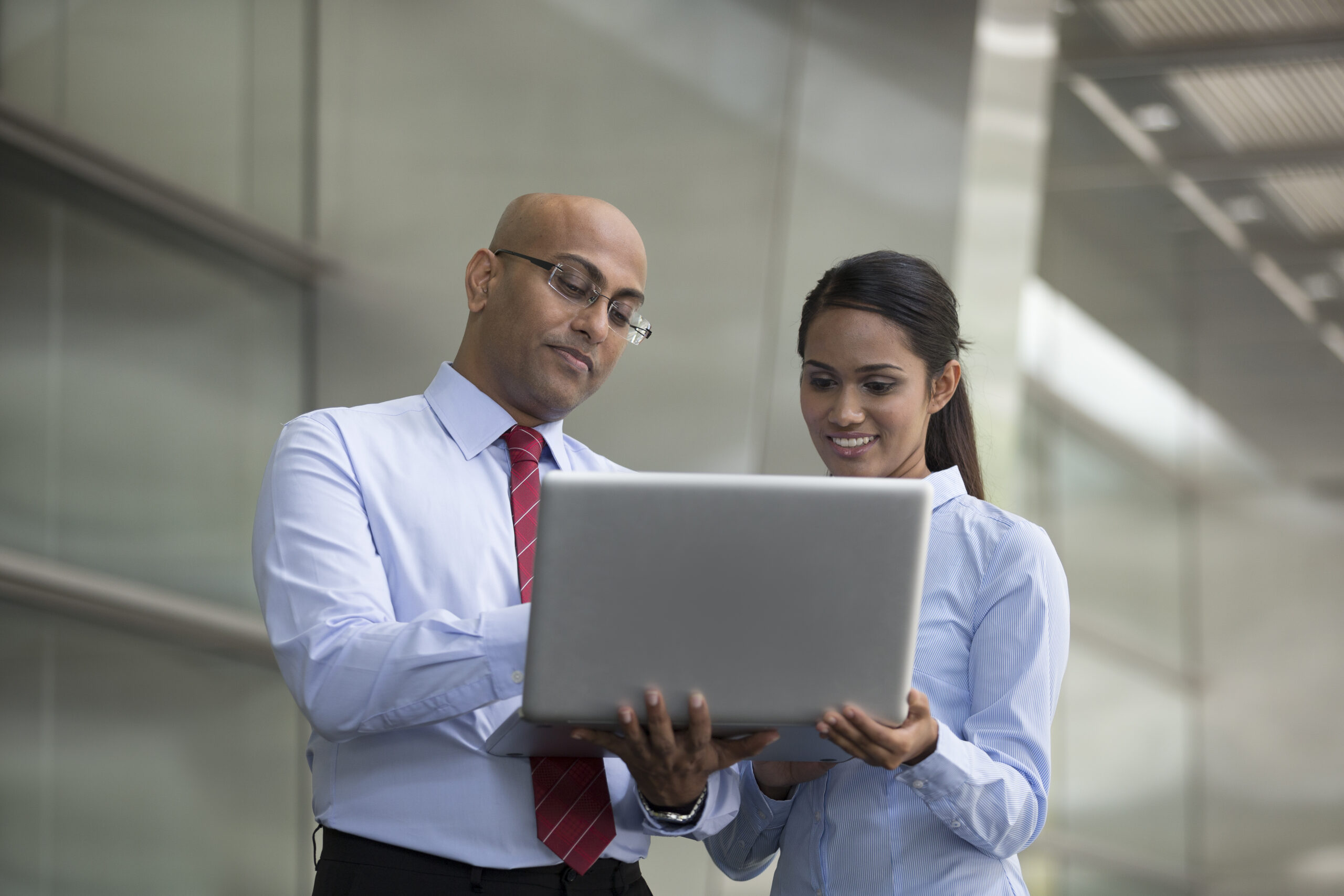 Eine indische Geschäftsfrau schaut gemeinsam mit einem indischen Geschäftsmann in ein Laptop. Im Hintergrund ein modernes Büro.
