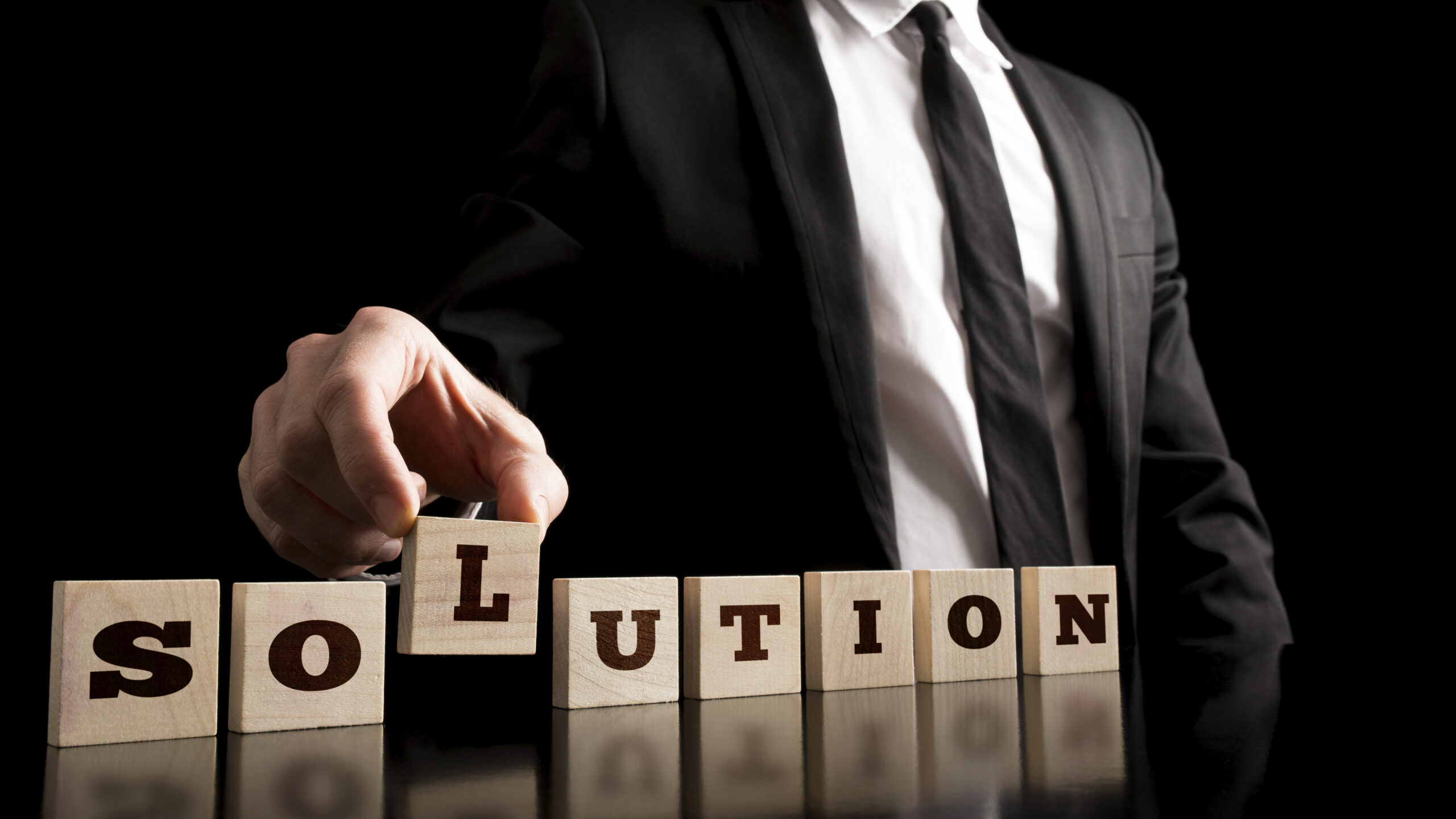 Die Buchstaben SOLUTION, einzeln auf Holzklötzchen gedruckt, stehen nebeneinander gereiht auf einem Tisch. Ein Mann im Businessoutfit steht dahinter und hält das Klötzchen mit dem Buchstaben L in seiner rechten Hand, so dass dieses leicht vom Tisch abgehoben ist.