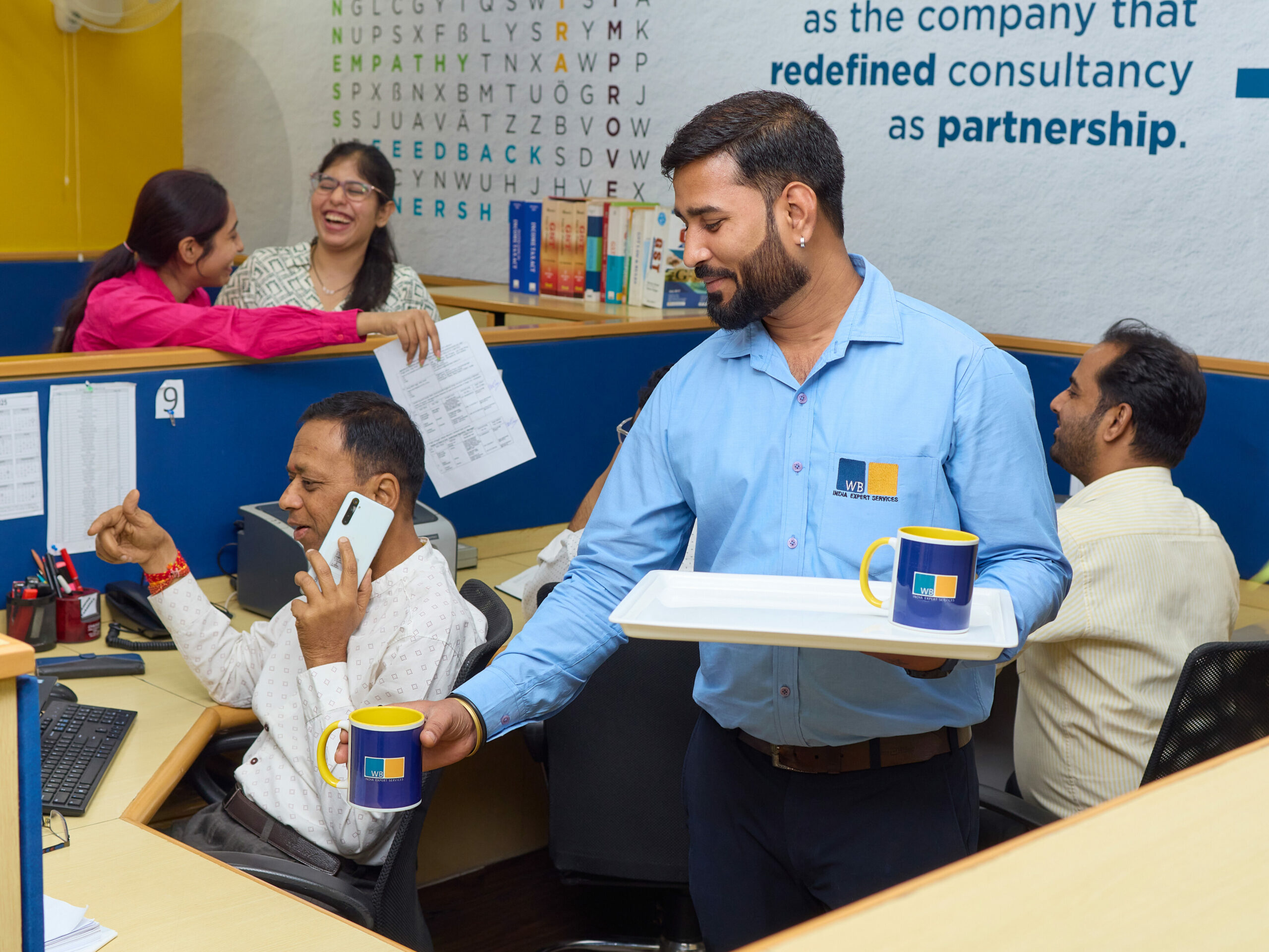 Gelöste Bürosituation im Wamser + Batra HQ in Delhi. Alle Mitarbeiter arbeiten, einer von ihnen serviert Heißgetränke in Bechern mit Firmenlogo.